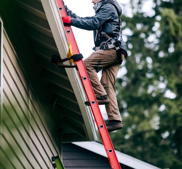 Worker on red ladder performing exterior home maintenance near roof.