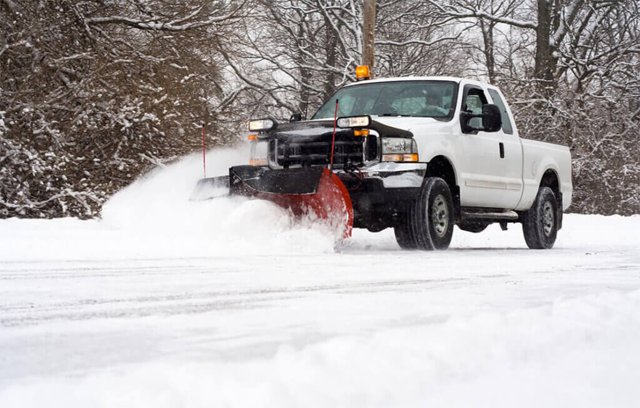 White truck with red plow clearing snow from a road; winter trees in background.