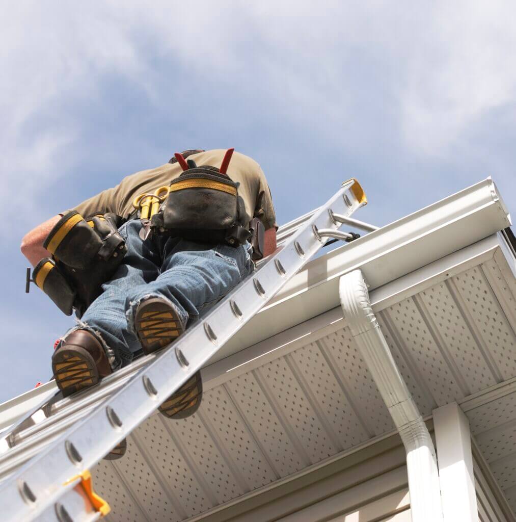 Person on a ladder wearing a tool belt, working near a home's white gutter and roof.
