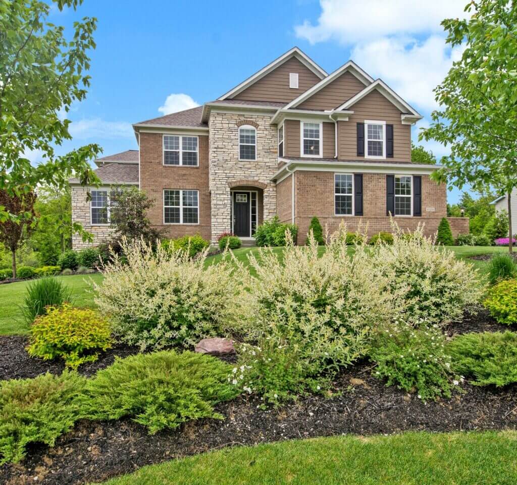 Brick and stone house with green shrubs and vibrant landscaping under a blue sky.