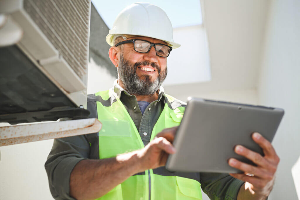 Worker in a hard hat and reflective vest using a tablet near an HVAC unit.