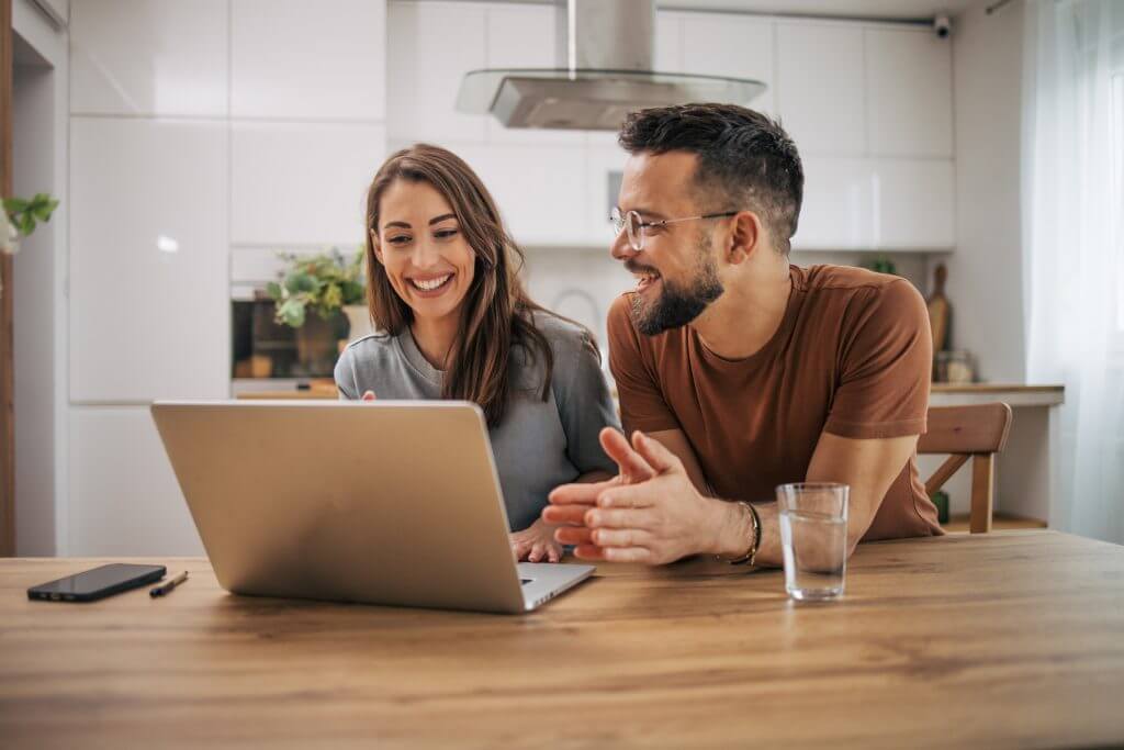 Smiling people viewing a silver laptop at a wooden table in a modern kitchen setting.