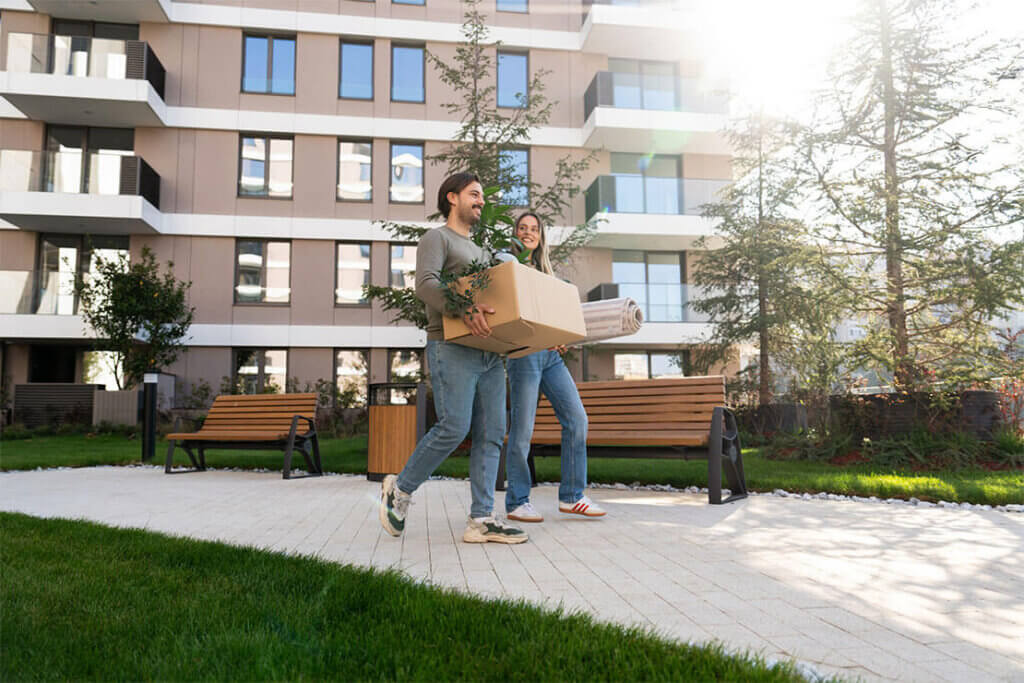 People carrying boxes in front of a modern commercial building with green landscaping.