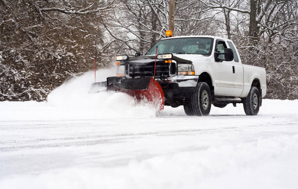 White truck with red plow clearing snow from a road; winter trees in background.
